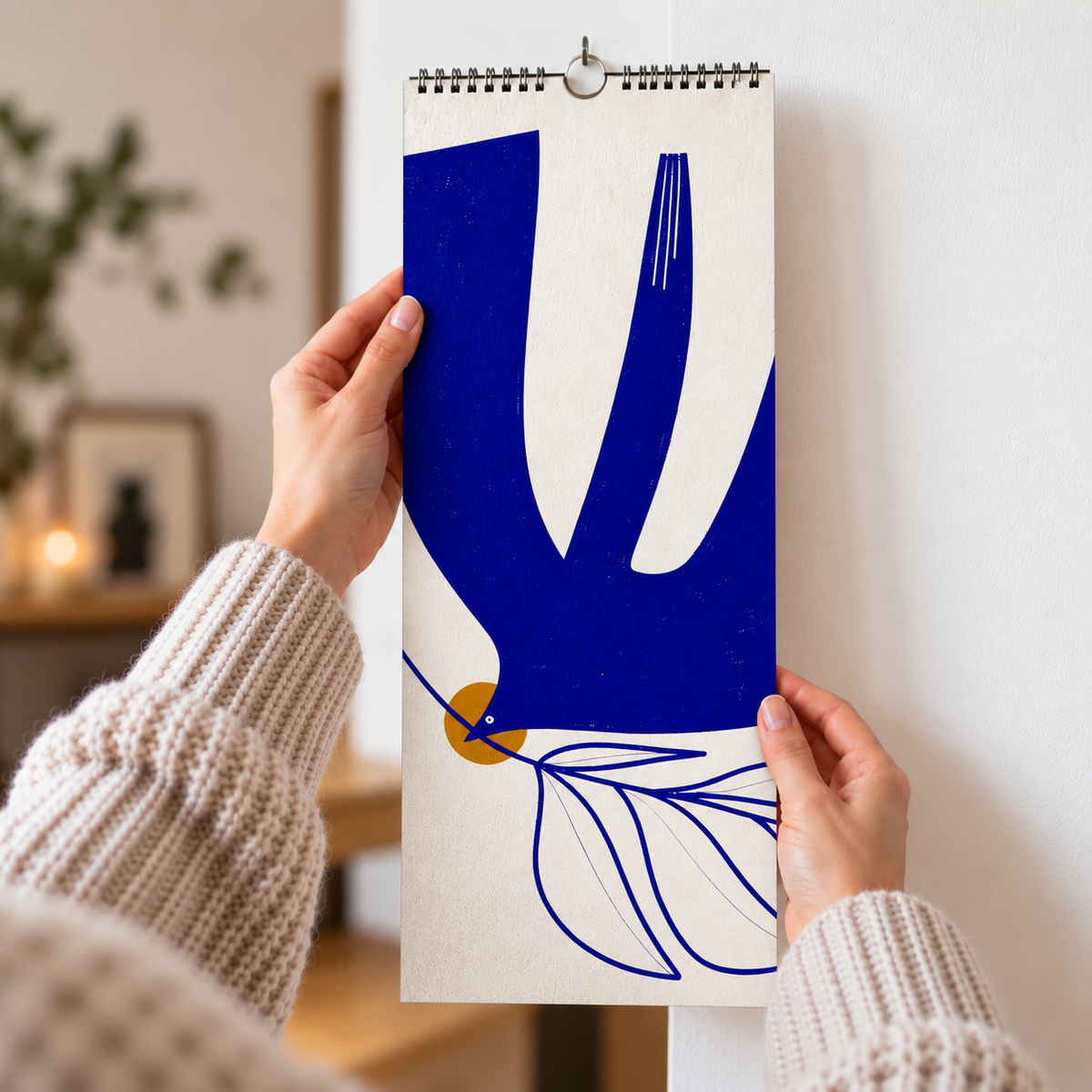 A person holds a spiral-bound wall calendar against a white wall. The calendar features a minimalist blue and white dove motif with a small mustard yellow circle and blue leaf line art.