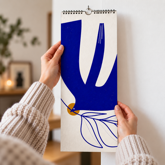 A person holds a spiral-bound wall calendar against a white wall. The calendar features a minimalist blue and white dove motif with a small mustard yellow circle and blue leaf line art.